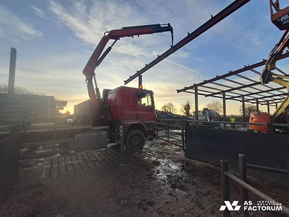 Red truck crane lifting a long steel beam on a construction site, with a steel frame behind