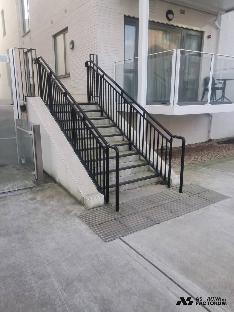 Outdoor concrete steps with black steel railings and tactile paving beside a white brick building
