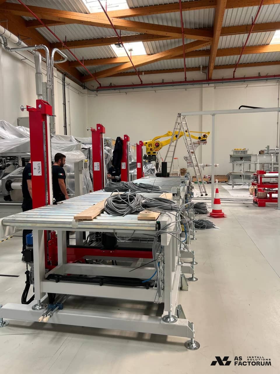 Roller conveyors with red lifting columns, workers and a yellow robot in the background