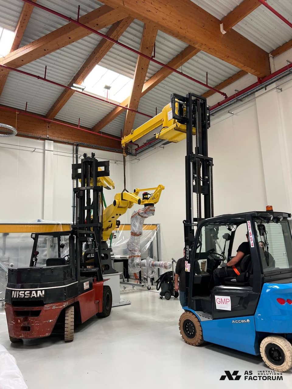 Wide shot of a yellow robot being moved by forklifts inside a hall