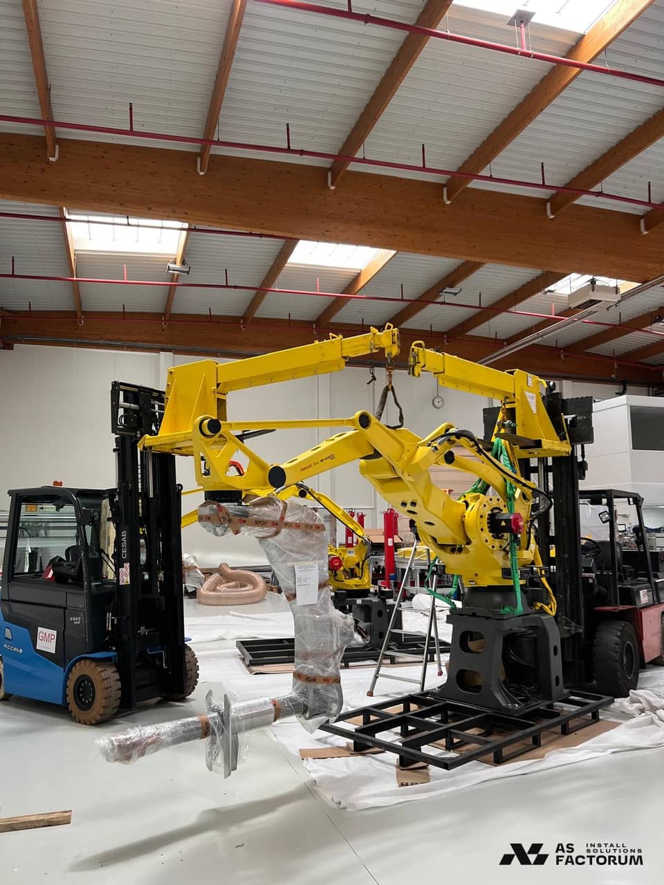 Yellow robot lifted by two forklifts, with its arm wrapped and lifting slings attached