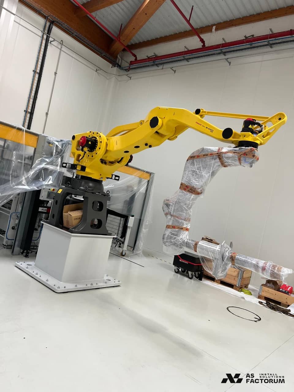 Yellow industrial robot on a pedestal in a hall, next to a second robot wrapped in film