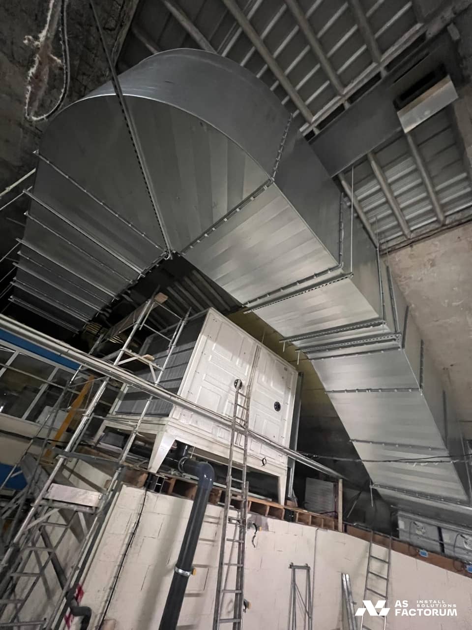 Large galvanised duct curving above a white fibreglass unit, with scaffolding alongside