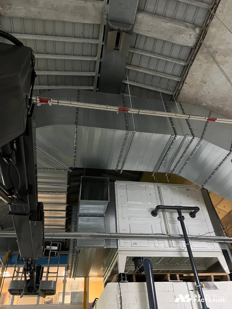 Wide galvanised duct bend with stiffening profiles above a white unit and a hydraulic boom arm
