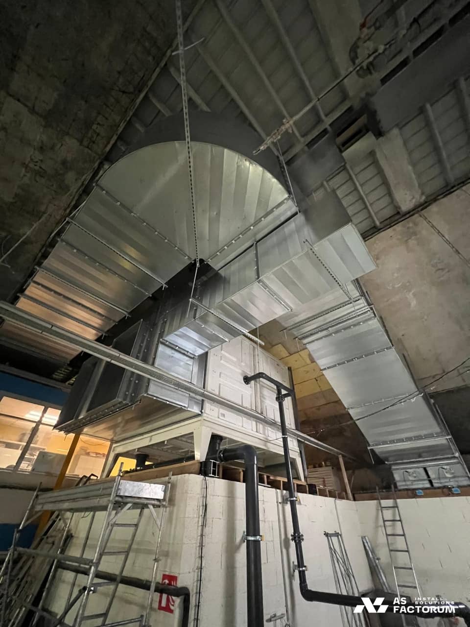 Curved galvanised duct elbows over a white unit, with grey pipes and scaffolding in the foreground