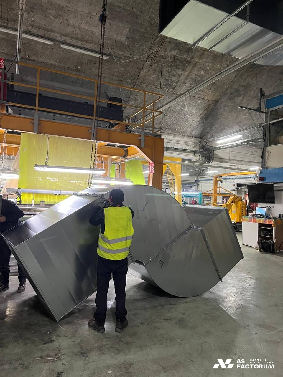 Two workers in hi-vis vests beside a large galvanised duct elbow on a textile hall floor